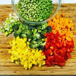 Raw vegetables diced up on a cutting board with a bowl of peas and beans.