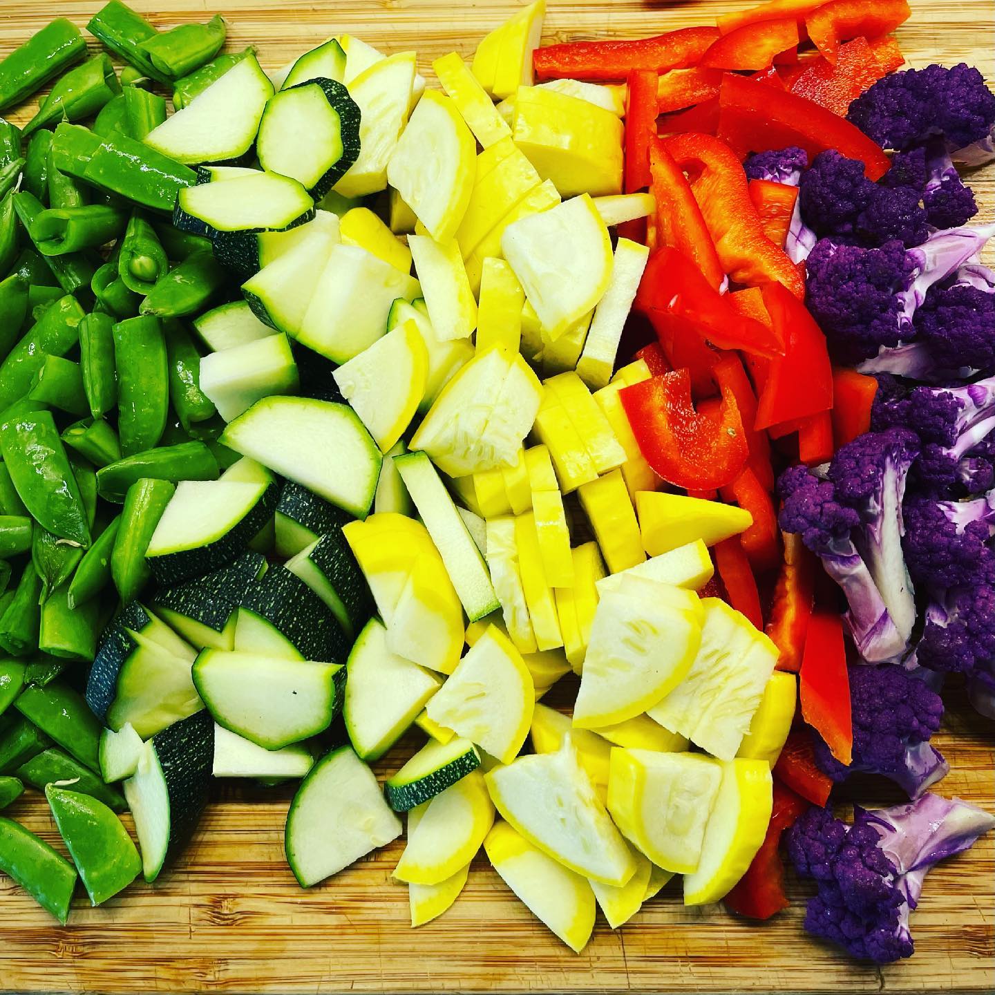 Summer vegetables cut up and placed in lines on a cutting board: green beans, zucchini, yellow squash, peppers and purple cauliflower.